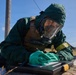 A person wearing full CBRN (Chemical, Biological, Radiological, Nuclear) protective gear, including a green hazmat suit, full-face gas mask, and green gloves, is writing on a document placed on a black case outdoors. The individual also wears a tan tactical vest over the protective suit. The setting appears to be a rural or field environment under clear blue skies, suggesting a military or emergency response exercise.