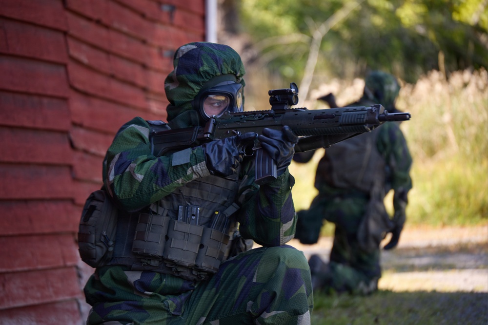 Two military personnel in full camouflage gear and gas masks are conducting what appears to be a tactical training exercise near a red brick building. The foreground soldier is in a kneeling position aiming a rifle equipped with a red dot sight, while a second soldier is visible in the background. The setting appears to be an outdoor urban or semi-rural training environment.