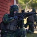 Two military personnel in full camouflage gear and gas masks are conducting what appears to be a tactical training exercise near a red brick building. The foreground soldier is in a kneeling position aiming a rifle equipped with a red dot sight, while a second soldier is visible in the background. The setting appears to be an outdoor urban or semi-rural training environment.