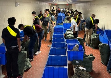 A long institutional corridor with red brick flooring is lined with blue plastic storage bins running down the center. A large group of individuals, most wearing black clothing with yellow reflective sashes/lanyards, are standing on both sides of the bins handling military-style duffel bags and camouflage gear. The setting and attire suggest a military induction, basic training intake, or ROTC/academy equipment issue process.