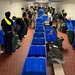 A long institutional corridor with red brick flooring is lined with blue plastic storage bins running down the center. A large group of individuals, most wearing black clothing with yellow reflective sashes/lanyards, are standing on both sides of the bins handling military-style duffel bags and camouflage gear. The setting and attire suggest a military induction, basic training intake, or ROTC/academy equipment issue process.