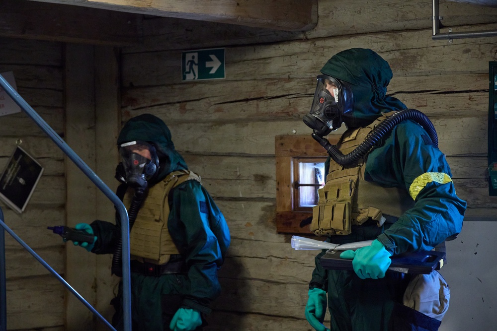 Two individuals wearing full CBRN (Chemical, Biological, Radiological, Nuclear) protective gear are shown inside a wooden-walled building. Both are equipped with gas masks, green hazmat suits, tactical vests, and green rubber gloves. They appear to be conducting an inspection or response operation, with one holding a detection device and documents.
