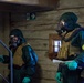 Two individuals wearing full CBRN (Chemical, Biological, Radiological, Nuclear) protective gear are shown inside a wooden-walled building. Both are equipped with gas masks, green hazmat suits, tactical vests, and green rubber gloves. They appear to be conducting an inspection or response operation, with one holding a detection device and documents.