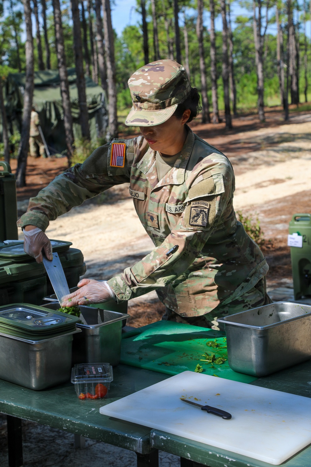 A U.S. Army soldier wearing Operational Camouflage Pattern (OCP) uniform with an Airborne tab and U.S. flag patch is preparing food at an outdoor field kitchen station. The soldier, whose name tape reads partially as '...EJO', is chopping vegetables on a green cutting board while wearing disposable gloves. The setting appears to be a military field training exercise in a wooded area.