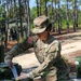 A U.S. Army soldier wearing Operational Camouflage Pattern (OCP) uniform with an Airborne tab and U.S. flag patch is preparing food at an outdoor field kitchen station. The soldier, whose name tape reads partially as '...EJO', is chopping vegetables on a green cutting board while wearing disposable gloves. The setting appears to be a military field training exercise in a wooded area.