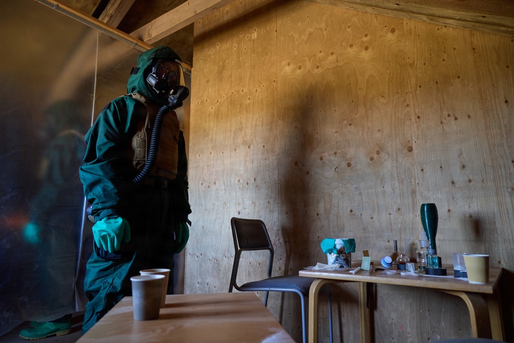 A person wearing a full green CBRN (Chemical, Biological, Radiological, Nuclear) protective suit with gas mask and green gloves stands inside a plywood-walled structure. The room contains tables with various small containers, cups, and laboratory-style glassware, suggesting a training or simulation scenario. The lighting is dim and dramatic, consistent with a military or emergency response exercise environment.