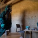 A person wearing a full green CBRN (Chemical, Biological, Radiological, Nuclear) protective suit with gas mask and green gloves stands inside a plywood-walled structure. The room contains tables with various small containers, cups, and laboratory-style glassware, suggesting a training or simulation scenario. The lighting is dim and dramatic, consistent with a military or emergency response exercise environment.