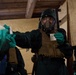 Personnel in full CBRN (Chemical, Biological, Radiological, Nuclear) protective gear are conducting what appears to be a training exercise or hazmat operation inside a wooden structure. The central figure wears a full-face gas mask, tactical vest, protective suit, and green chemical-resistant gloves, and is handling small paper or card items. A second similarly equipped individual is visible on the left, and a third partially visible on the right.