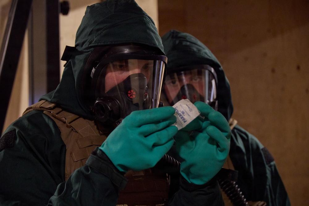 Two individuals wearing full CBRN (Chemical, Biological, Radiological, Nuclear) protective gear are examining a small packaged item. Both are equipped with full-face gas masks, green protective hoods, green nitrile/rubber gloves, and tactical vests. The foreground individual is closely inspecting what appears to be a labeled test kit or detection packet.