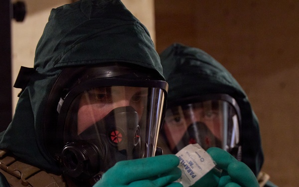 Two individuals wearing full CBRN (Chemical, Biological, Radiological, Nuclear) protective gear are examining a small packaged item. Both are equipped with full-face gas masks, green protective hoods, green nitrile/rubber gloves, and tactical vests. The foreground individual is closely inspecting what appears to be a labeled test kit or detection packet.