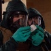 Two individuals wearing full CBRN (Chemical, Biological, Radiological, Nuclear) protective gear are examining a small packaged item. Both are equipped with full-face gas masks, green protective hoods, green nitrile/rubber gloves, and tactical vests. The foreground individual is closely inspecting what appears to be a labeled test kit or detection packet.