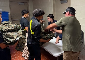 A group of young people, likely students, are gathered around a folding table in what appears to be a school or institutional hallway. An adult in a green shirt and khaki pants is distributing or inspecting military camouflage uniforms (MARPAT pattern) to the students. This appears to be a JROTC or similar military youth program uniform issue event.