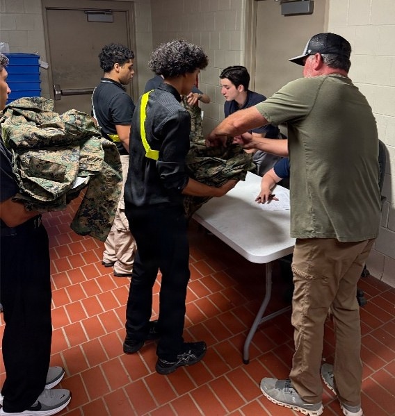 A group of young people, likely students, are gathered around a folding table in what appears to be a school or institutional hallway. An adult in a green shirt and khaki pants is distributing or inspecting military camouflage uniforms (MARPAT pattern) to the students. This appears to be a JROTC or similar military youth program uniform issue event.