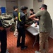 A group of young people, likely students, are gathered around a folding table in what appears to be a school or institutional hallway. An adult in a green shirt and khaki pants is distributing or inspecting military camouflage uniforms (MARPAT pattern) to the students. This appears to be a JROTC or similar military youth program uniform issue event.