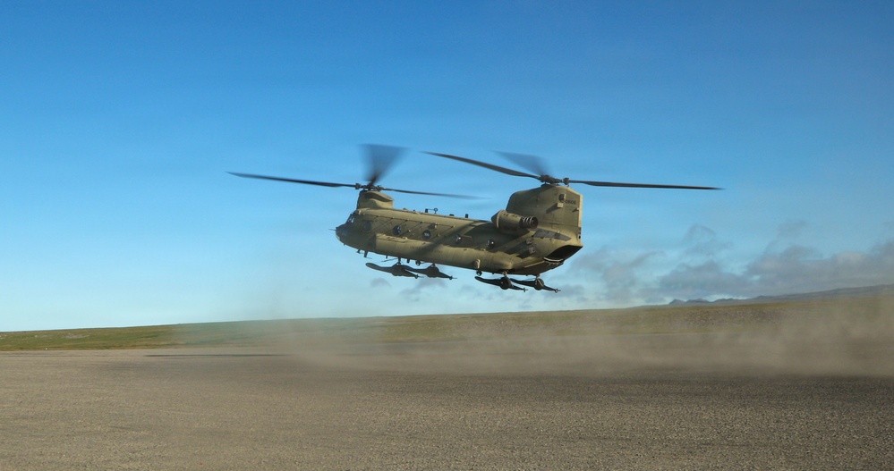 A military tandem-rotor helicopter, consistent with a CH-47 Chinook, is captured in low-level flight or during takeoff/landing over a flat, arid terrain. The rotors are visibly spinning, creating motion blur, and the downwash is kicking up a significant dust cloud to the rear of the aircraft. The sky is clear and blue, suggesting daytime operations in an open, semi-arid environment.
