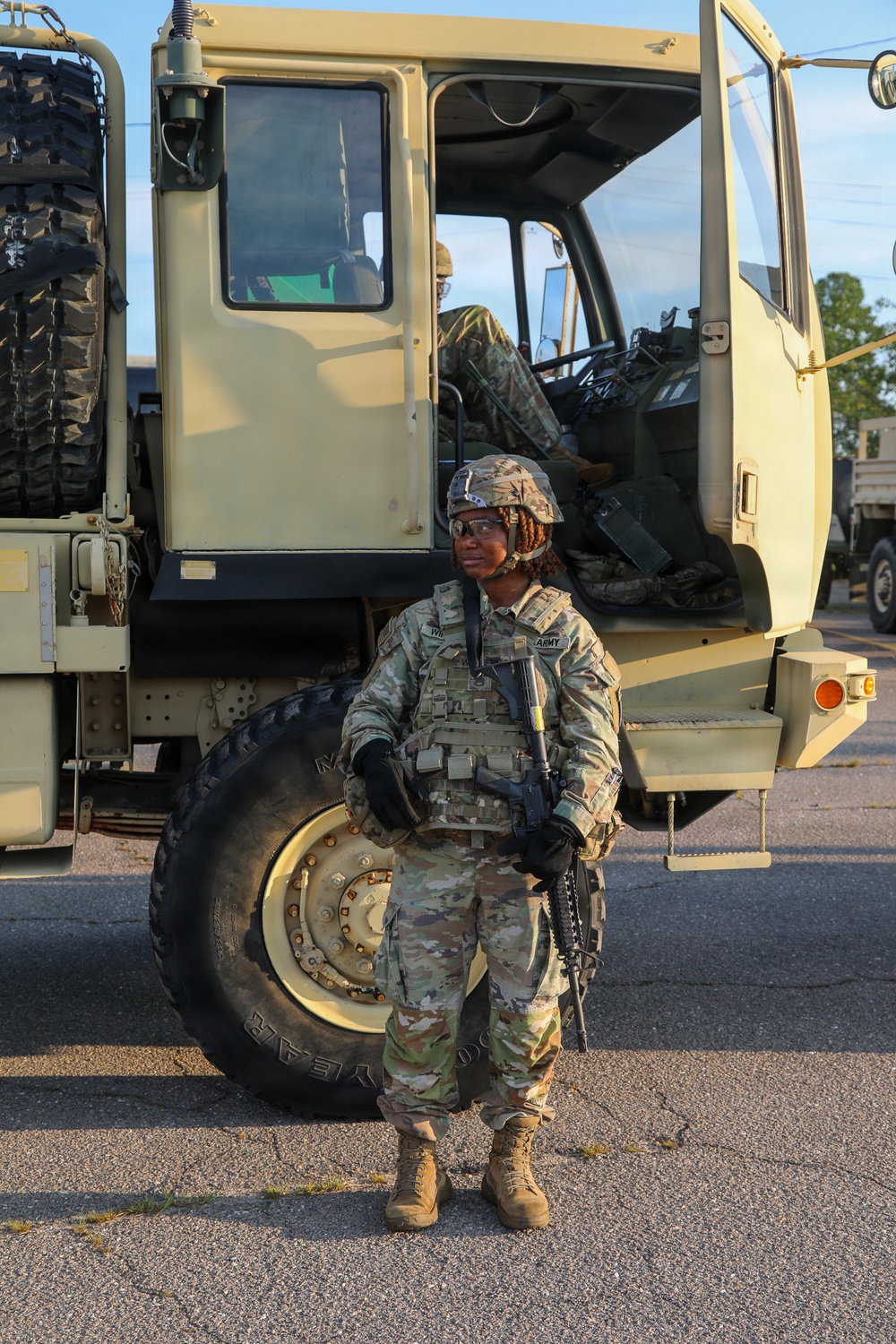 A U.S. Army soldier in full combat gear stands at parade rest in front of a large military tactical truck (likely an LMTV or FMTV). The soldier is wearing OCP (Operational Camouflage Pattern) uniform, body armor, ACH helmet, and is holding an M4 carbine rifle. A second soldier is partially visible seated inside the open cab of the vehicle.