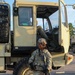 A U.S. Army soldier in full combat gear stands at parade rest in front of a large military tactical truck (likely an LMTV or FMTV). The soldier is wearing OCP (Operational Camouflage Pattern) uniform, body armor, ACH helmet, and is holding an M4 carbine rifle. A second soldier is partially visible seated inside the open cab of the vehicle.