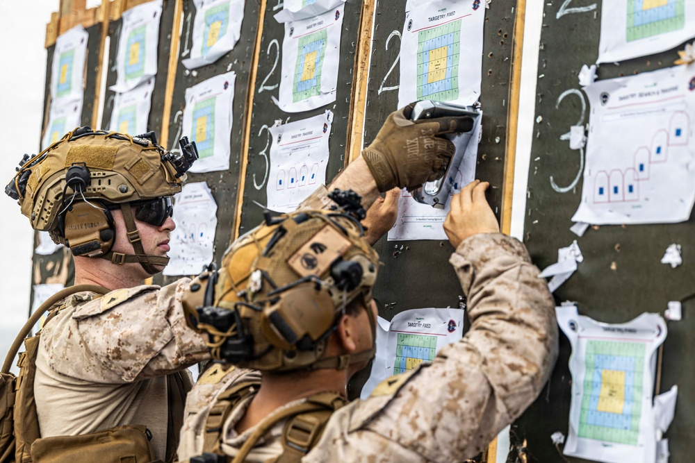Two military personnel in desert camouflage uniforms and tactical helmets are examining and posting documents on a large dark board. The board contains multiple printed target sheets with grid patterns in blue, yellow, and green colors, along with chalked lane numbers. This appears to be a firing range scoring or target assessment activity.