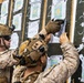 Two military personnel in desert camouflage uniforms and tactical helmets are examining and posting documents on a large dark board. The board contains multiple printed target sheets with grid patterns in blue, yellow, and green colors, along with chalked lane numbers. This appears to be a firing range scoring or target assessment activity.