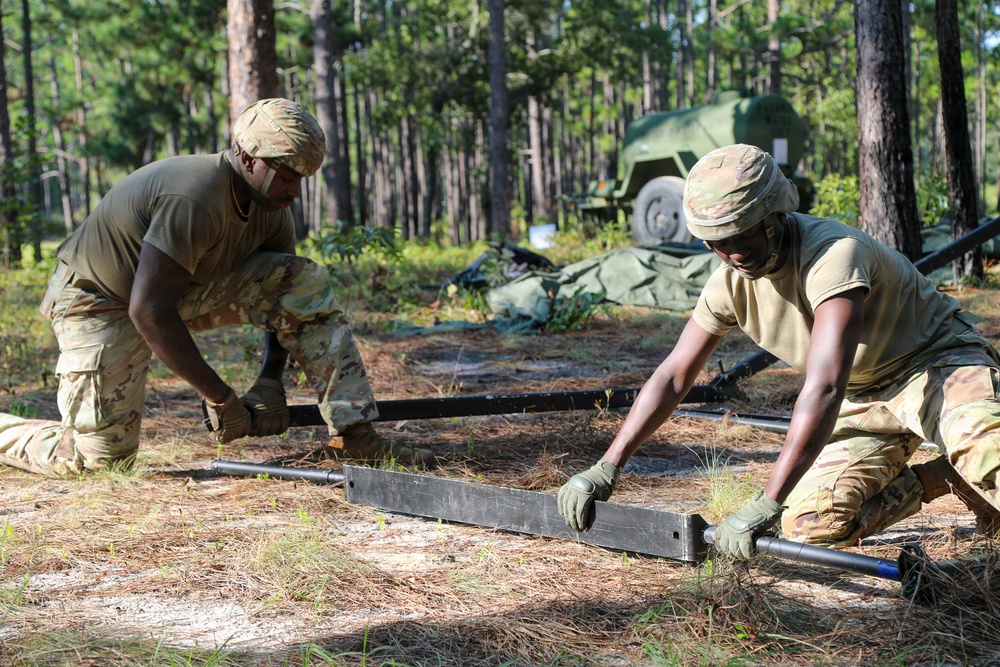 Two military personnel in camouflage uniforms are working together to assemble or disassemble metal frame components on a sandy, pine needle-covered ground in a forested area. Both soldiers are wearing multicam pattern uniforms, patrol caps, and tactical gloves. A military vehicle and green canvas equipment are visible in the background.