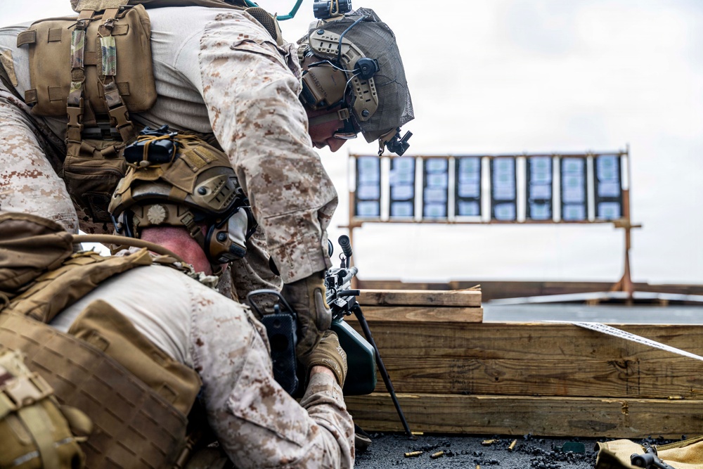 Two military personnel in desert MARPAT camouflage uniforms are positioned behind a wooden barricade on what appears to be a shooting range. One service member is leaning over to assist the other, who is in a prone or low position with a rifle equipped with a bipod. A target board is visible in the background at range distance.