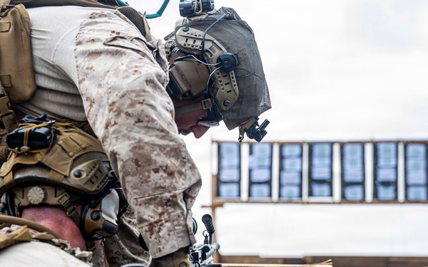 Two military personnel in desert MARPAT camouflage uniforms are positioned behind a wooden barricade on what appears to be a shooting range. One service member is leaning over to assist the other, who is in a prone or low position with a rifle equipped with a bipod. A target board is visible in the background at range distance.