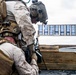 Two military personnel in desert MARPAT camouflage uniforms are positioned behind a wooden barricade on what appears to be a shooting range. One service member is leaning over to assist the other, who is in a prone or low position with a rifle equipped with a bipod. A target board is visible in the background at range distance.