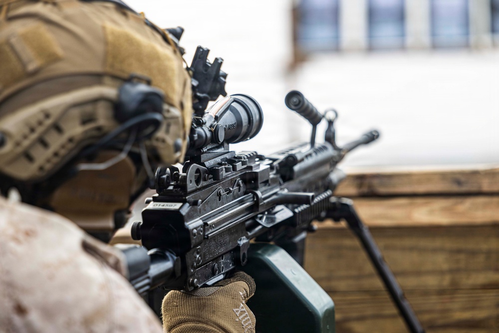 A military personnel in desert camouflage uniform and tactical helmet is positioned behind a wooden barrier, aiming a light machine gun. The weapon appears to be an M249 SAW (Squad Automatic Weapon) equipped with an optic sight, bipod deployed on the wooden surface, and an ammunition box attached. The image is taken from a close rear-angle perspective, emphasizing the weapon system and operator's gear.