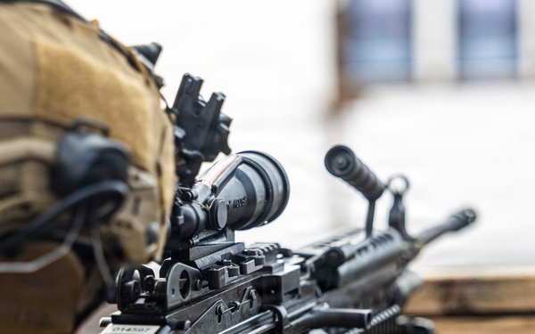 A military personnel in desert camouflage uniform and tactical helmet is positioned behind a wooden barrier, aiming a light machine gun. The weapon appears to be an M249 SAW (Squad Automatic Weapon) equipped with an optic sight, bipod deployed on the wooden surface, and an ammunition box attached. The image is taken from a close rear-angle perspective, emphasizing the weapon system and operator's gear.
