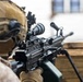 A military personnel in desert camouflage uniform and tactical helmet is positioned behind a wooden barrier, aiming a light machine gun. The weapon appears to be an M249 SAW (Squad Automatic Weapon) equipped with an optic sight, bipod deployed on the wooden surface, and an ammunition box attached. The image is taken from a close rear-angle perspective, emphasizing the weapon system and operator's gear.