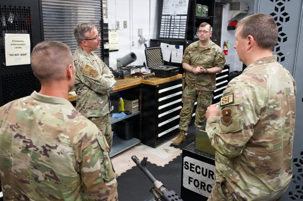 Four military personnel in OCP (Operational Camouflage Pattern) uniforms are gathered in what appears to be a Security Forces armory or weapons maintenance room. They appear to be engaged in a briefing or discussion, with one senior member addressing the others. The setting includes weapon storage, maintenance equipment, and Security Forces signage.