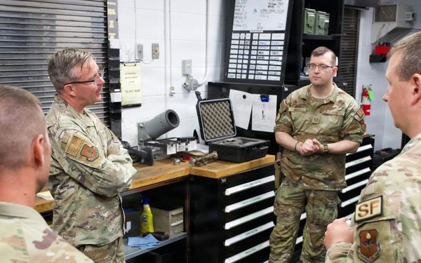 Four military personnel in OCP (Operational Camouflage Pattern) uniforms are gathered in what appears to be a Security Forces armory or weapons maintenance room. They appear to be engaged in a briefing or discussion, with one senior member addressing the others. The setting includes weapon storage, maintenance equipment, and Security Forces signage.