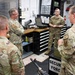Four military personnel in OCP (Operational Camouflage Pattern) uniforms are gathered in what appears to be a Security Forces armory or weapons maintenance room. They appear to be engaged in a briefing or discussion, with one senior member addressing the others. The setting includes weapon storage, maintenance equipment, and Security Forces signage.