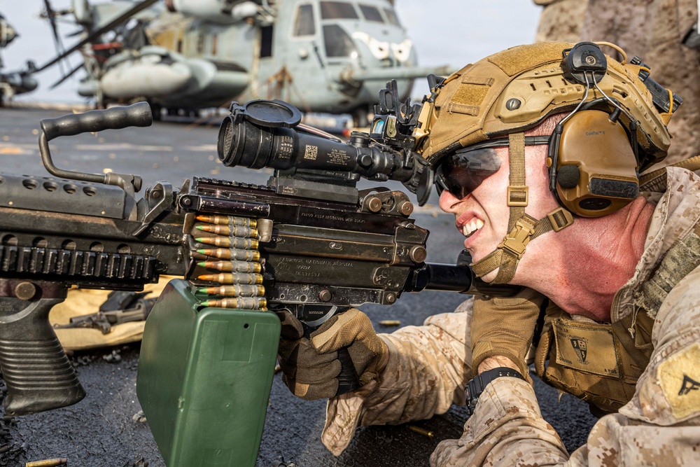A U.S. Marine in full combat gear is operating a belt-fed machine gun, likely an M249 SAW or M240, in a prone firing position on what appears to be the deck of a naval vessel. The Marine is wearing MARPAT desert camouflage, a tan FAST helmet with accessories, hearing protection, and tactical gloves. Military helicopters, consistent with CH-53 Sea Stallions, are visible in the background.