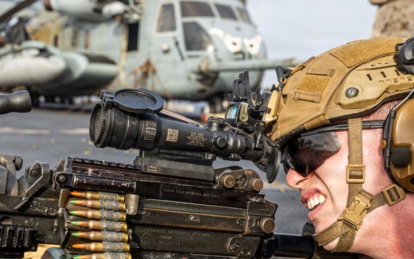 A U.S. Marine in full combat gear is operating a belt-fed machine gun, likely an M249 SAW or M240, in a prone firing position on what appears to be the deck of a naval vessel. The Marine is wearing MARPAT desert camouflage, a tan FAST helmet with accessories, hearing protection, and tactical gloves. Military helicopters, consistent with CH-53 Sea Stallions, are visible in the background.