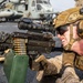 A U.S. Marine in full combat gear is operating a belt-fed machine gun, likely an M249 SAW or M240, in a prone firing position on what appears to be the deck of a naval vessel. The Marine is wearing MARPAT desert camouflage, a tan FAST helmet with accessories, hearing protection, and tactical gloves. Military helicopters, consistent with CH-53 Sea Stallions, are visible in the background.