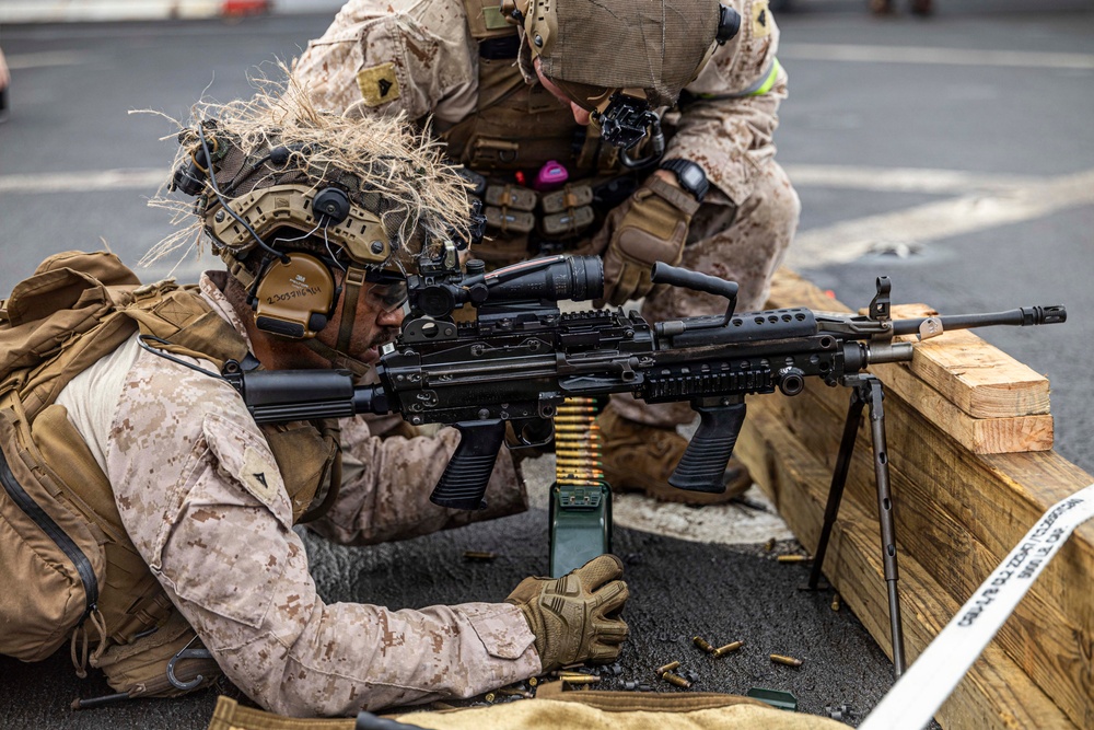 Two military personnel in MARPAT desert camouflage uniforms are conducting a live-fire training exercise. The prone shooter is operating what appears to be an M249 Squad Automatic Weapon (SAW) mounted on a bipod, with a belt-fed ammunition box visible. A second service member is positioned behind in a support/observer role.