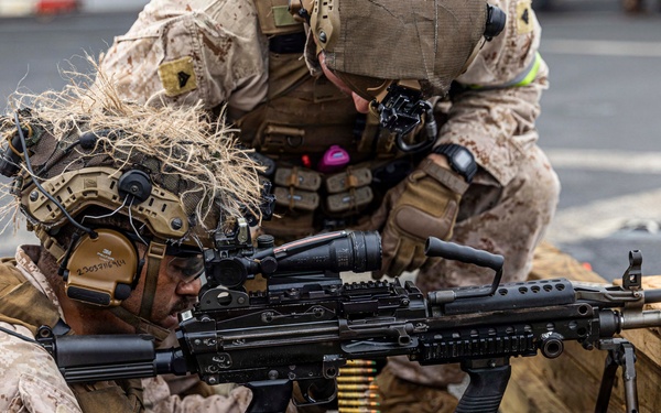 Two military personnel in MARPAT desert camouflage uniforms are conducting a live-fire training exercise. The prone shooter is operating what appears to be an M249 Squad Automatic Weapon (SAW) mounted on a bipod, with a belt-fed ammunition box visible. A second service member is positioned behind in a support/observer role.