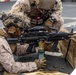Two military personnel in MARPAT desert camouflage uniforms are conducting a live-fire training exercise. The prone shooter is operating what appears to be an M249 Squad Automatic Weapon (SAW) mounted on a bipod, with a belt-fed ammunition box visible. A second service member is positioned behind in a support/observer role.