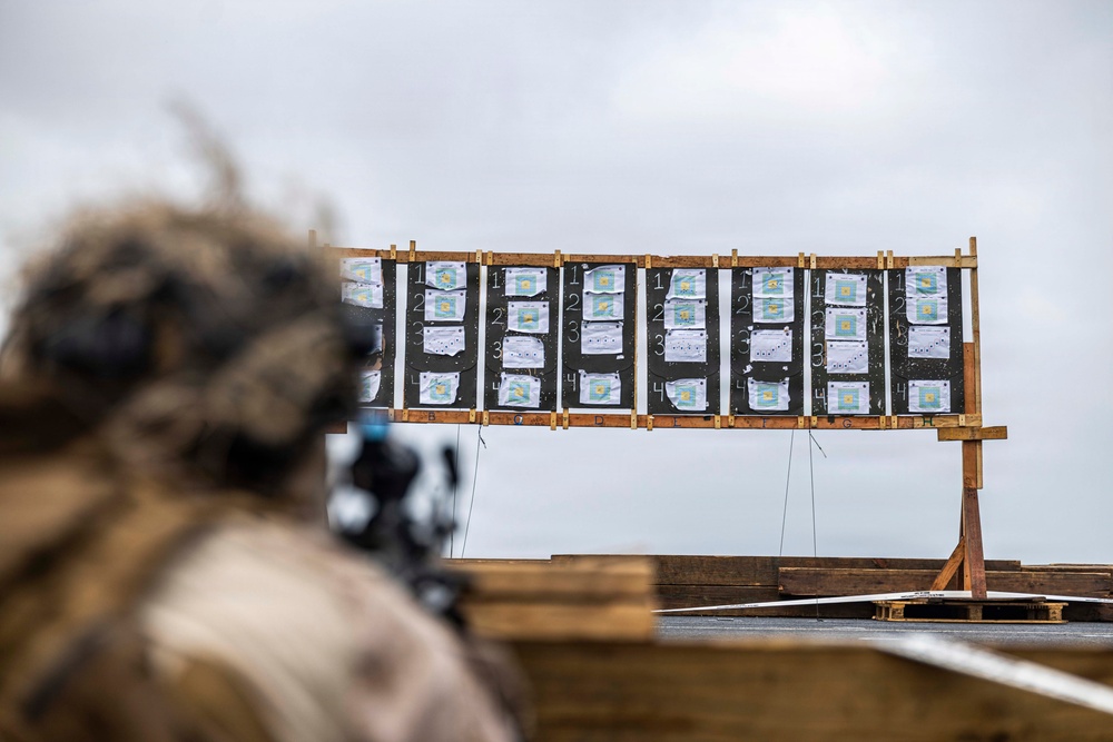 A shooter in camouflage gear and helmet is aiming a scoped rifle at a wooden target board downrange. The target board displays multiple paper shooting targets arranged in columns, showing numbered scoring zones with visible bullet impact marks. The setting appears to be an outdoor military or law enforcement firing range.