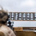 A shooter in camouflage gear and helmet is aiming a scoped rifle at a wooden target board downrange. The target board displays multiple paper shooting targets arranged in columns, showing numbered scoring zones with visible bullet impact marks. The setting appears to be an outdoor military or law enforcement firing range.