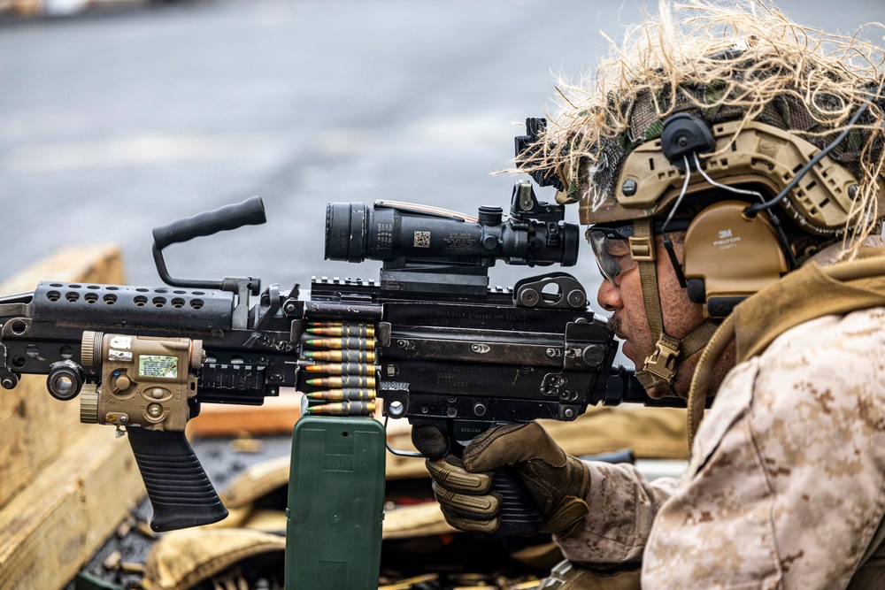 A military service member in full combat gear is aiming a belt-fed machine gun, likely an M249 SAW or M240, equipped with an optical scope and laser targeting device. The operator is wearing MARPAT camouflage uniform, a tan tactical helmet with ghillie netting, hearing protection, and tactical gloves. The image appears to depict a live-fire training exercise or operational readiness drill.
