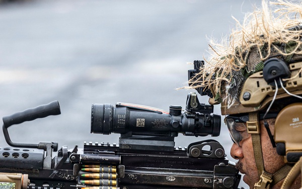A military service member in full combat gear is aiming a belt-fed machine gun, likely an M249 SAW or M240, equipped with an optical scope and laser targeting device. The operator is wearing MARPAT camouflage uniform, a tan tactical helmet with ghillie netting, hearing protection, and tactical gloves. The image appears to depict a live-fire training exercise or operational readiness drill.