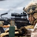 A military service member in full combat gear is aiming a belt-fed machine gun, likely an M249 SAW or M240, equipped with an optical scope and laser targeting device. The operator is wearing MARPAT camouflage uniform, a tan tactical helmet with ghillie netting, hearing protection, and tactical gloves. The image appears to depict a live-fire training exercise or operational readiness drill.