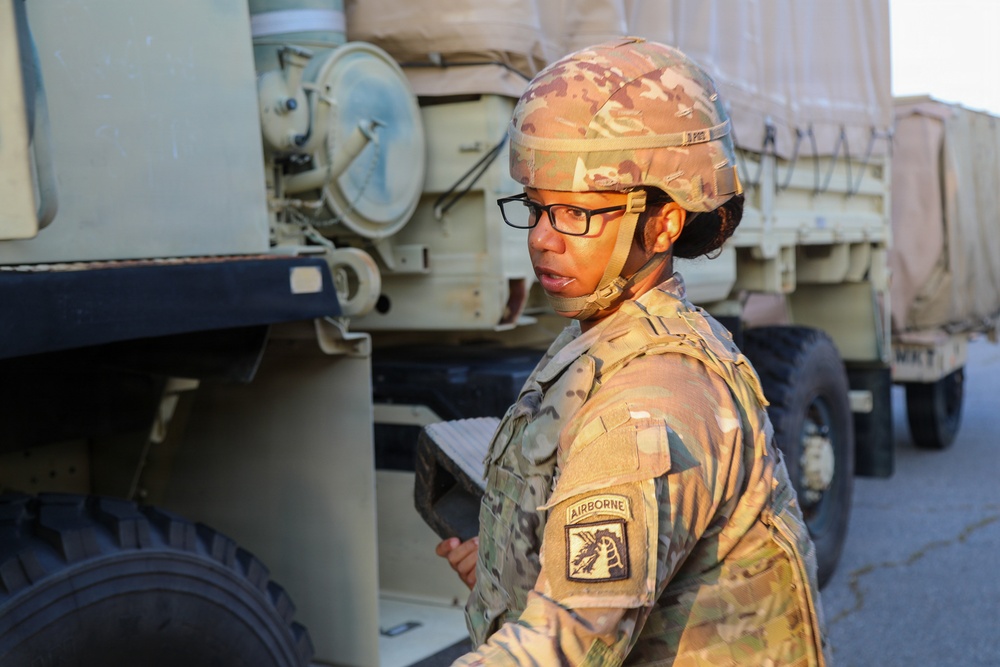 A female U.S. Army soldier wearing Operational Camouflage Pattern (OCP) uniform and combat helmet is conducting what appears to be an inspection or inventory check near military vehicles. She is holding a clipboard or tablet and wearing glasses, suggesting an administrative or logistical role. The setting appears to be a motor pool or staging area with tan military trucks visible in the background.