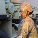 A female U.S. Army soldier wearing Operational Camouflage Pattern (OCP) uniform and combat helmet is conducting what appears to be an inspection or inventory check near military vehicles. She is holding a clipboard or tablet and wearing glasses, suggesting an administrative or logistical role. The setting appears to be a motor pool or staging area with tan military trucks visible in the background.