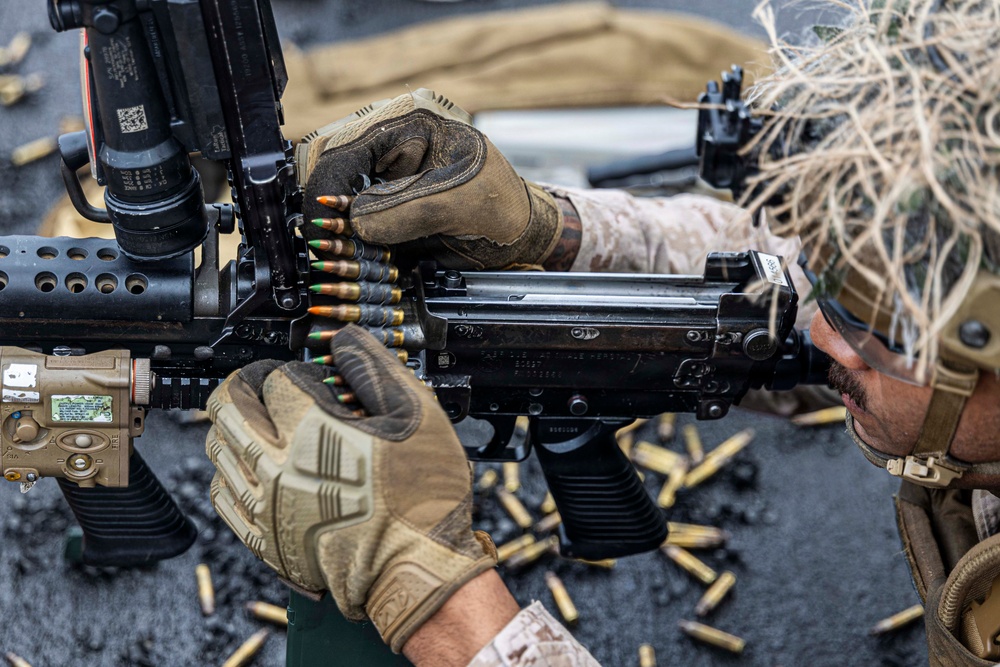 Two military personnel wearing desert camouflage uniforms and tactical gloves are loading a belt-fed machine gun. Spent brass casings are scattered on the ground surface below the weapon, indicating recent firing activity. The weapon appears to be an M249 or similar belt-fed light machine gun with various tactical accessories mounted.