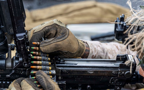 Two military personnel wearing desert camouflage uniforms and tactical gloves are loading a belt-fed machine gun. Spent brass casings are scattered on the ground surface below the weapon, indicating recent firing activity. The weapon appears to be an M249 or similar belt-fed light machine gun with various tactical accessories mounted.