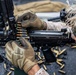 Two military personnel wearing desert camouflage uniforms and tactical gloves are loading a belt-fed machine gun. Spent brass casings are scattered on the ground surface below the weapon, indicating recent firing activity. The weapon appears to be an M249 or similar belt-fed light machine gun with various tactical accessories mounted.