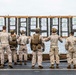 A group of approximately seven military personnel in desert camouflage uniforms are standing on what appears to be a ship's deck, reviewing shooting target results posted on a large wooden board. The targets display numbered scoring zones and shot grouping patterns, suggesting a marksmanship qualification or training exercise. The overcast sky and visible ocean in the background confirm an at-sea naval vessel setting.